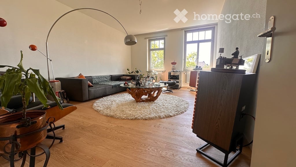A modern living room with a grey sectional sofa, a round coffee table, a floor lamp, and a potted plant. The room features large windows, wooden flooring, and a cozy white rug.