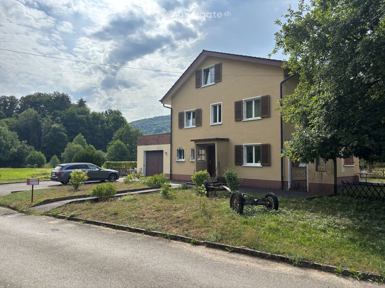 Two-story house with beige exterior, multiple windows with shutters, and a small garden area with a parked car nearby.