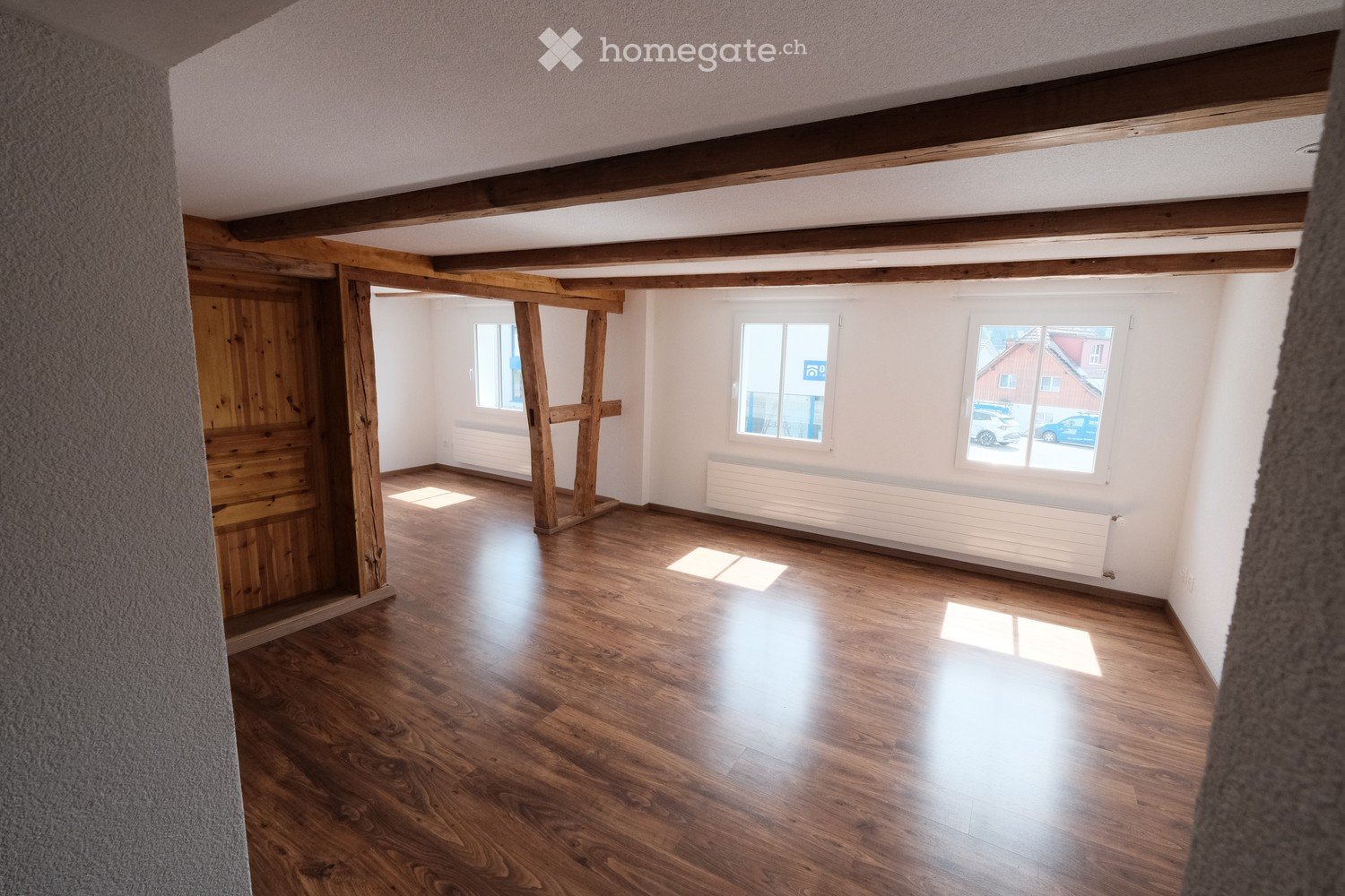 Empty room with wooden flooring, white walls, exposed wooden beams, and large windows letting in natural light.