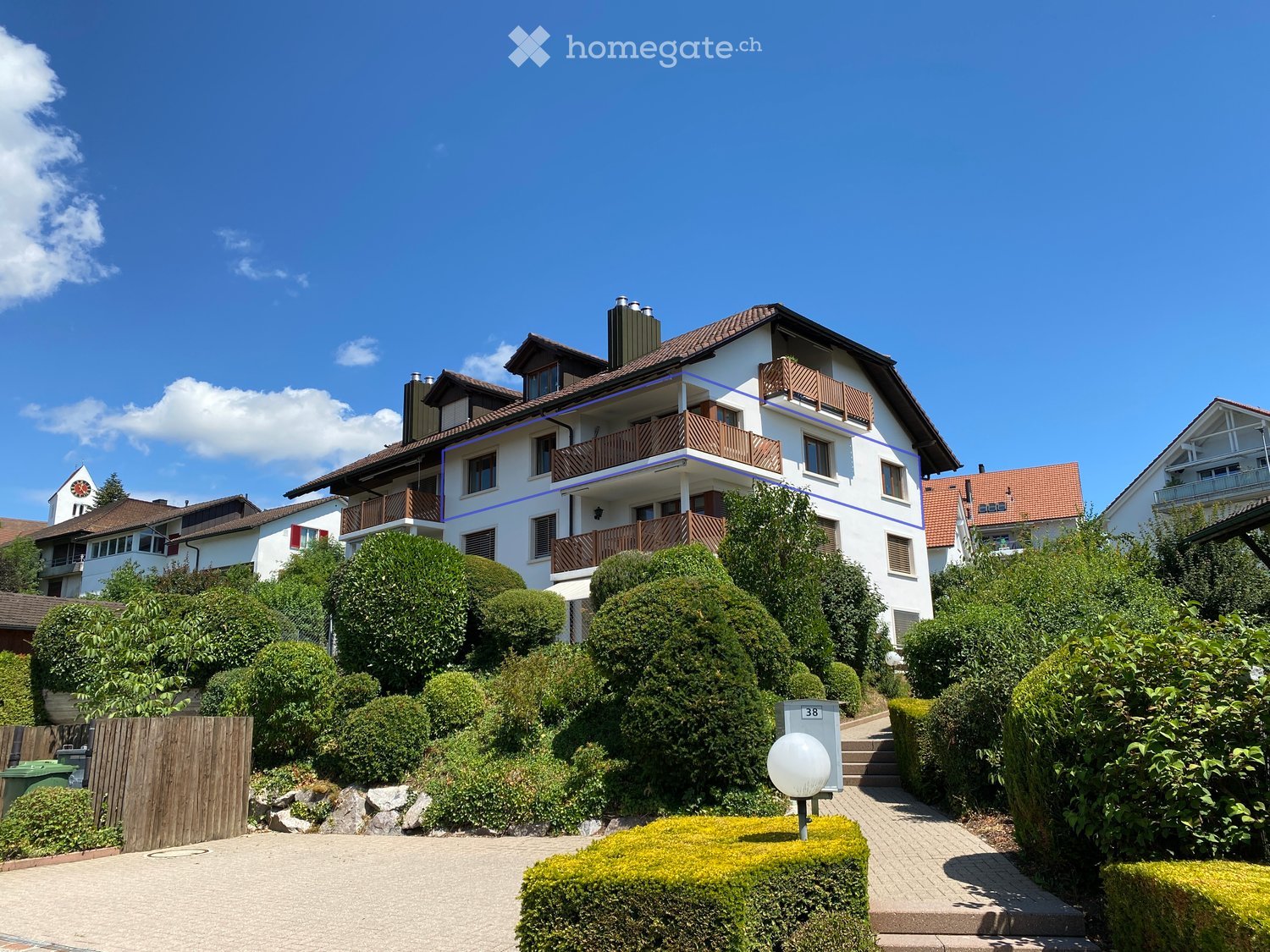 A two-story house with white walls, brown roof, multiple windows, and balconies, surrounded by well-maintained greenery and shrubs.