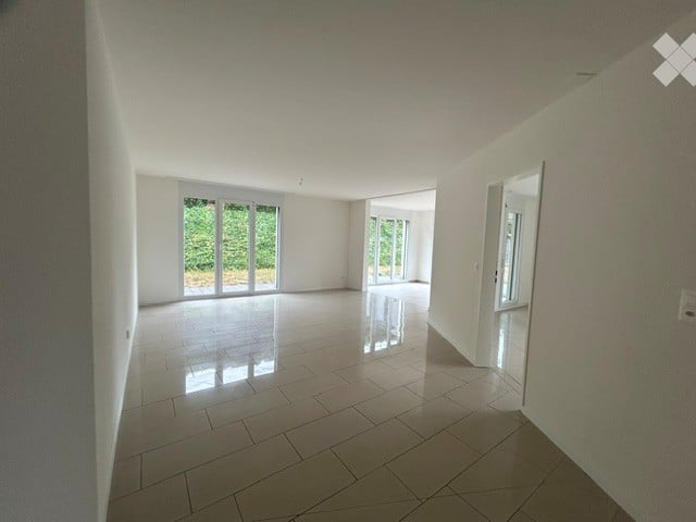 Empty room with white walls, tiled floor, large sliding glass doors leading to an outdoor area, and a visible doorway to another room.