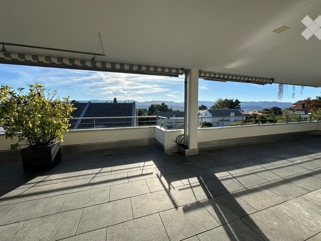 Spacious balcony with tiled flooring, white railings, and a potted plant, offering a scenic view of the surrounding area and mountains.