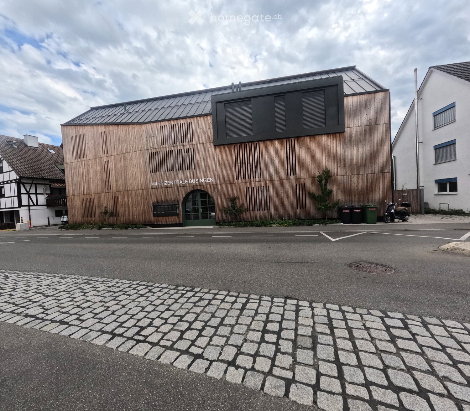 Modern building with wooden facade and large windows, featuring a sign reading "MILCHZENTRALE BUSINGEN" and a scooter parked nearby.