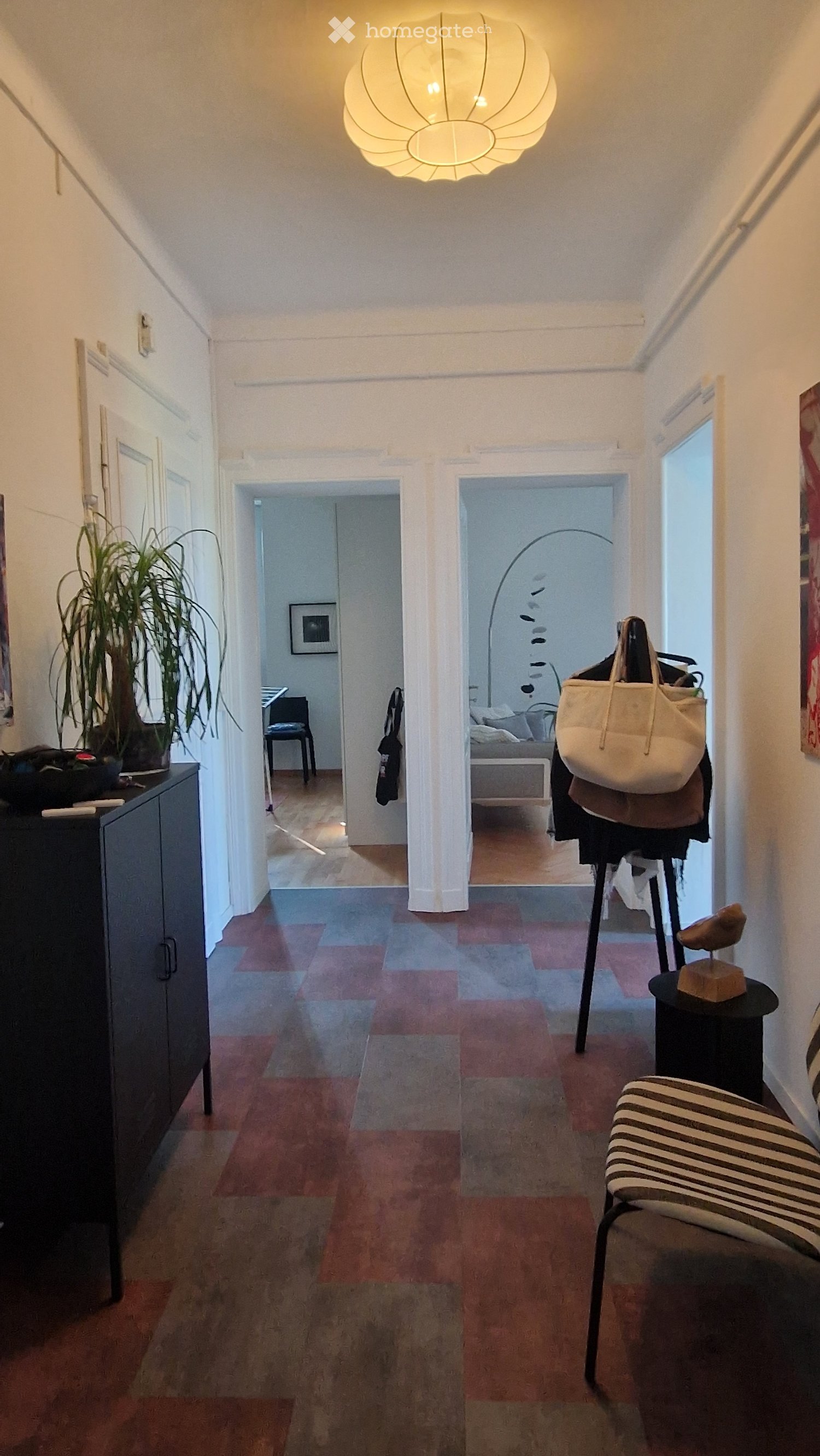 Hallway with wooden flooring, white walls, and a hanging light fixture. Visible rooms include a bedroom and a living area with a chair and plant.