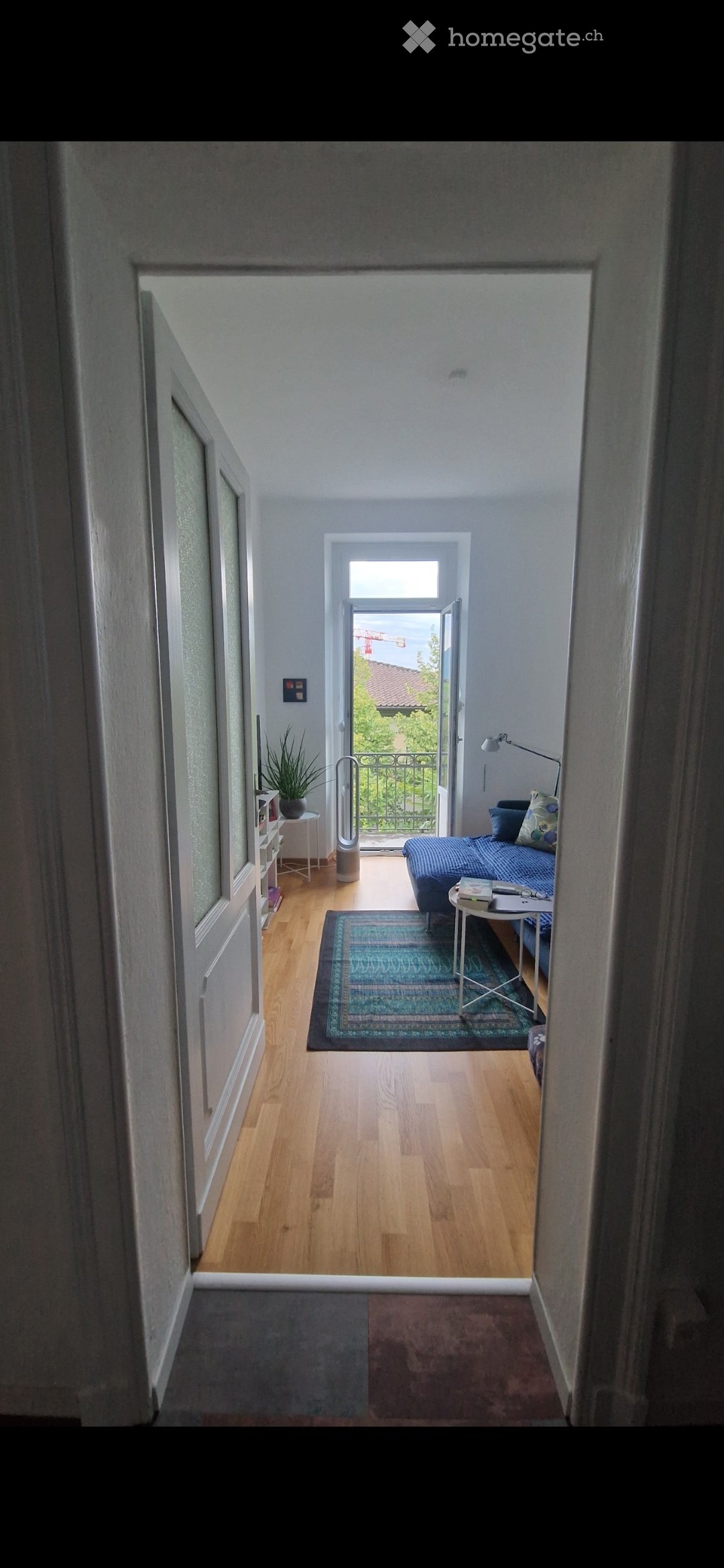 A modern, well-lit bedroom with wooden flooring, a blue sofa, a small table with a lamp, a rug, and a plant near the balcony entrance.