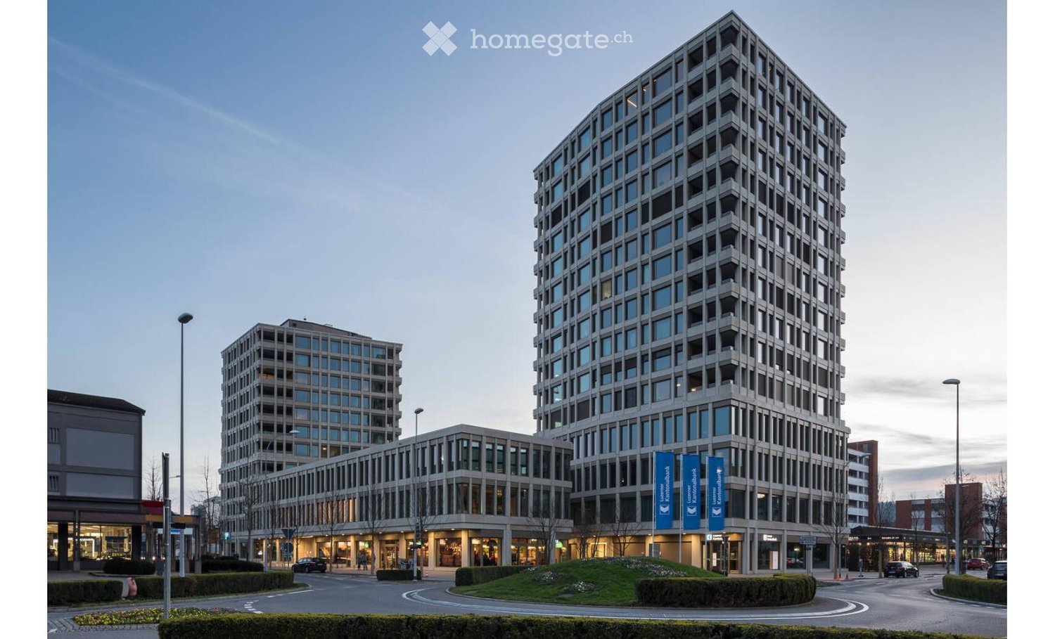 Modern multi-story building with a geometric facade, large windows, and surrounding landscaped area with pathways and streetlights.