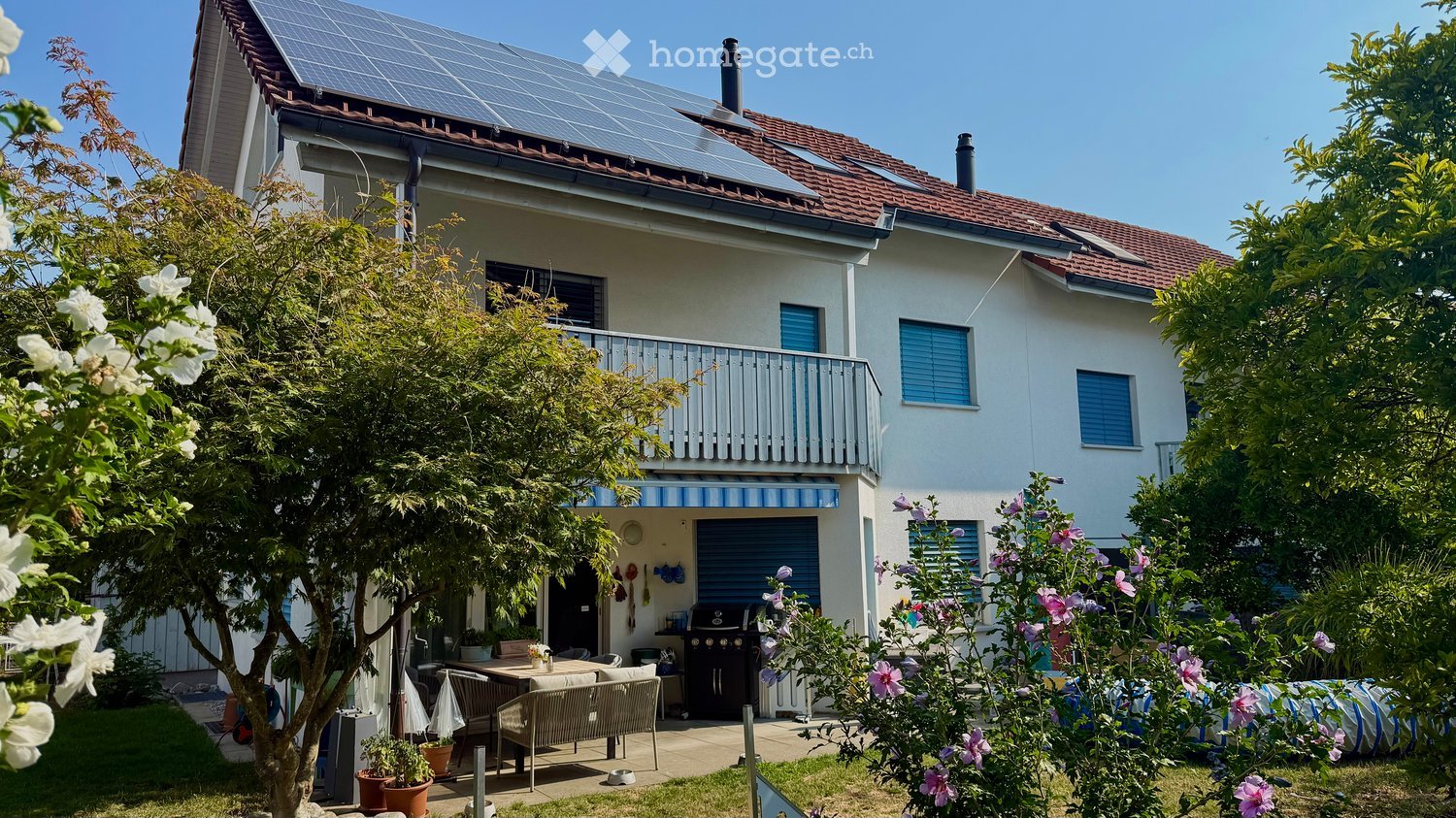 Two-story house with solar panels on the roof, a balcony, and a well-maintained garden with outdoor seating and a barbecue.