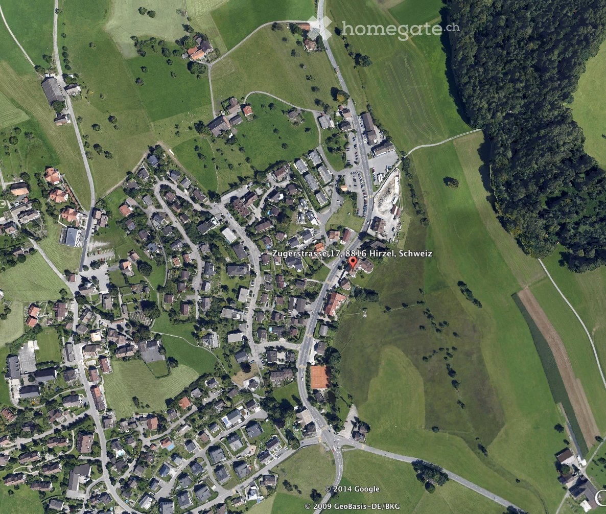 Aerial view of a rural village with scattered houses, green fields, and roads. The area includes residential buildings, agricultural land, and a small cluster of structures near a road intersection.