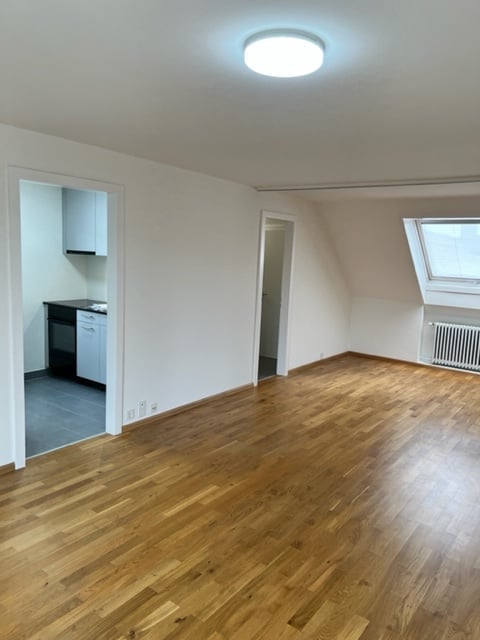 Empty room with wooden flooring, white walls, and a small kitchen area visible through an open doorway. The space is well-lit with a ceiling light fixture.