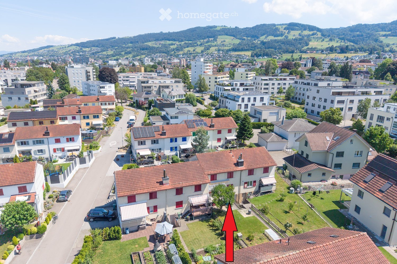Aerial view of a residential neighborhood with multiple houses, gardens, and a scenic backdrop of hills and trees.