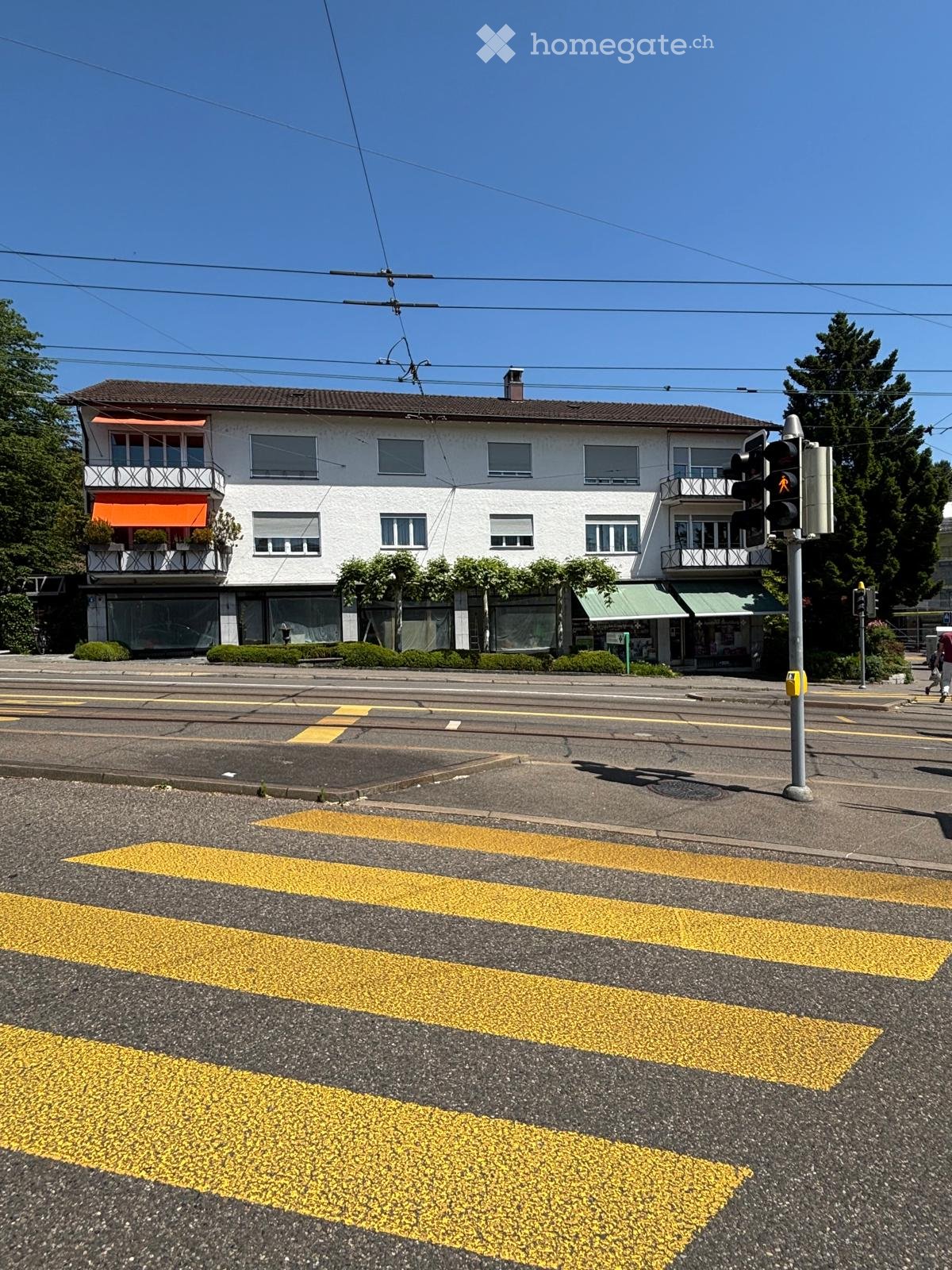 Two-story white building with multiple windows, balconies, and awnings, located at a street corner with a pedestrian crossing.