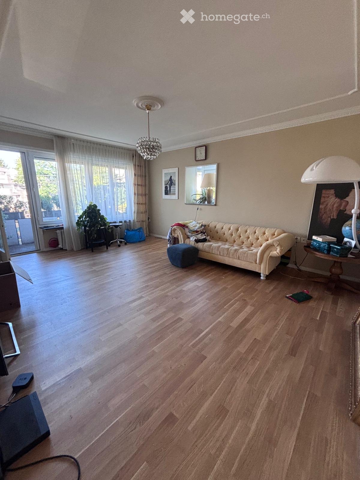 Living room with wooden flooring, a cream-colored sofa, ottoman, side table, lamp, mirror, picture frame, and a plant near the window.