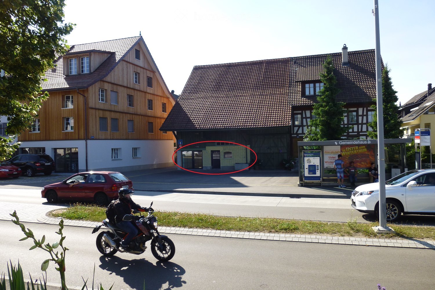 Two-story residential buildings with pitched roofs, multiple windows, and a small garage. A motorcycle rider is passing by on the road.