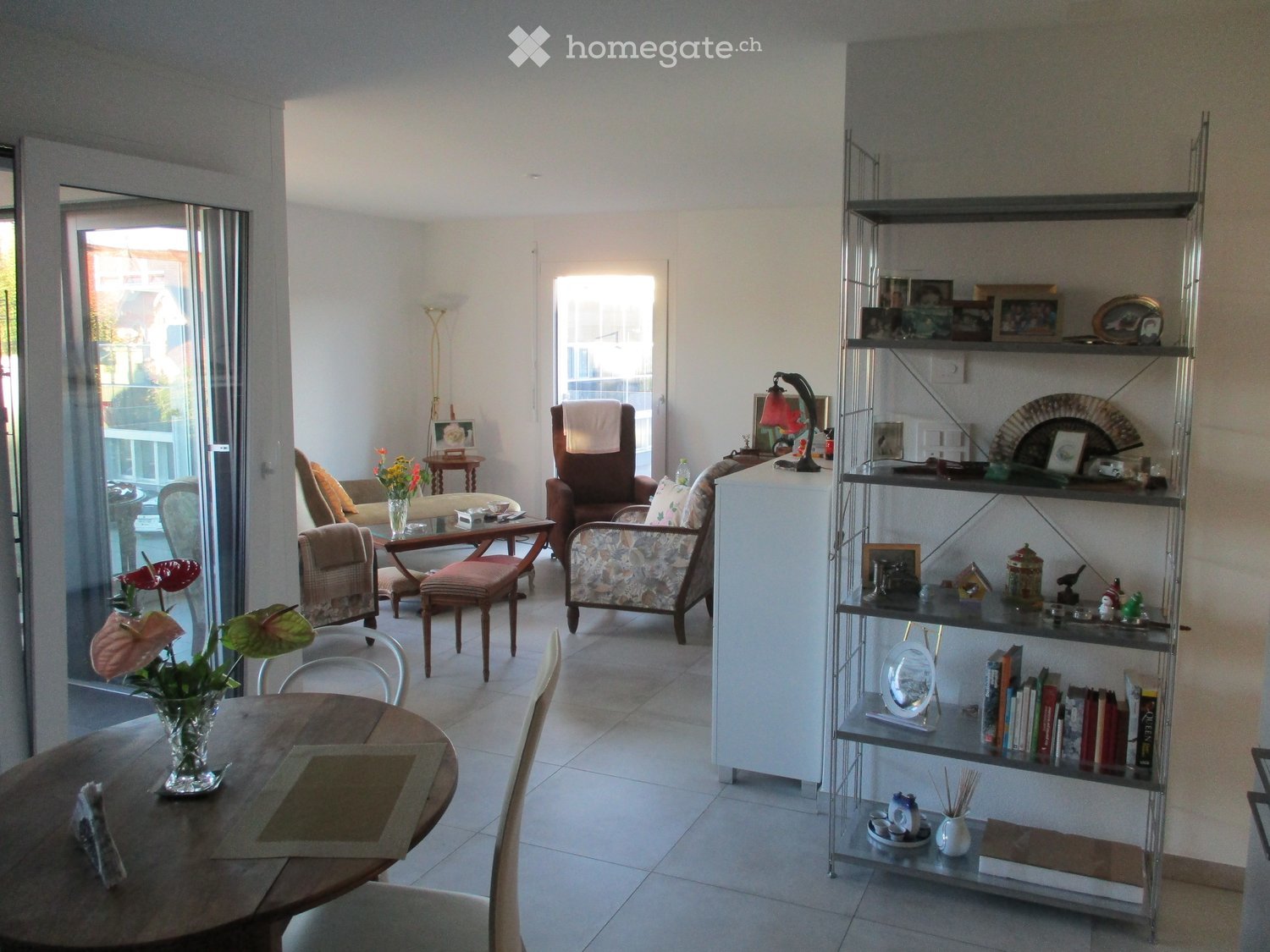 Bright living room with tiled floor, large windows, and sliding glass doors leading to a balcony. Features a round wooden dining table with chairs, a sofa, armchairs, a bookshelf with decorative items, and a lamp.