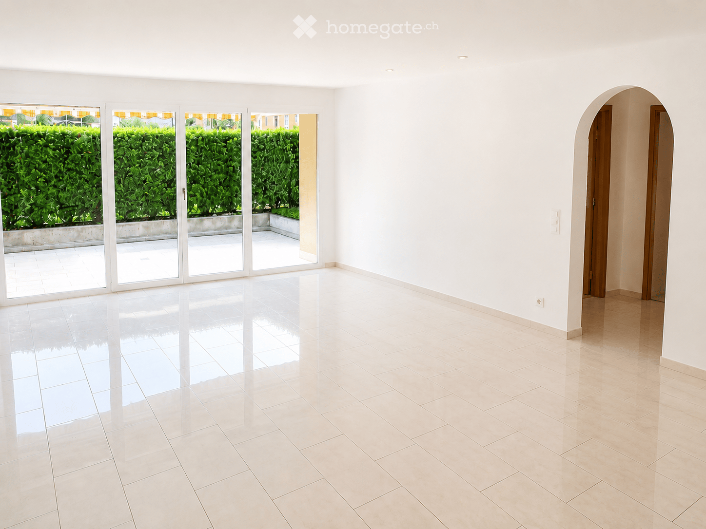 Empty room with white walls, tiled floors, large sliding glass doors leading to a patio, and an arched doorway.