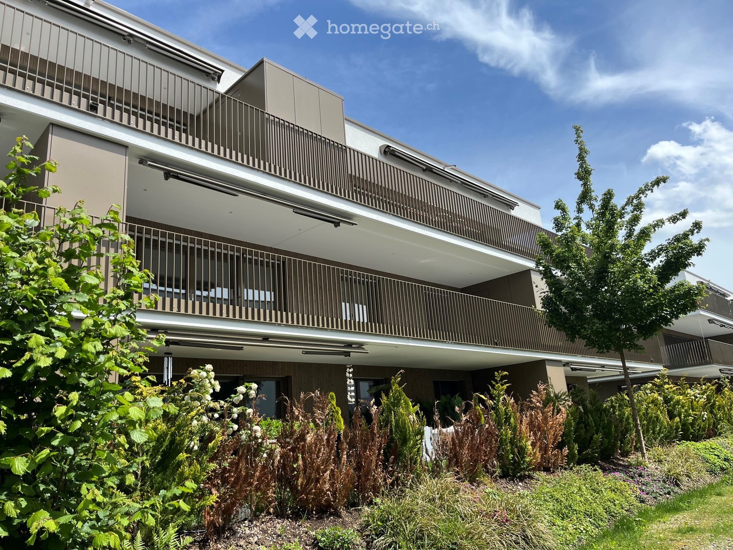 Modern multi-story building with balconies, surrounded by lush greenery and trees.