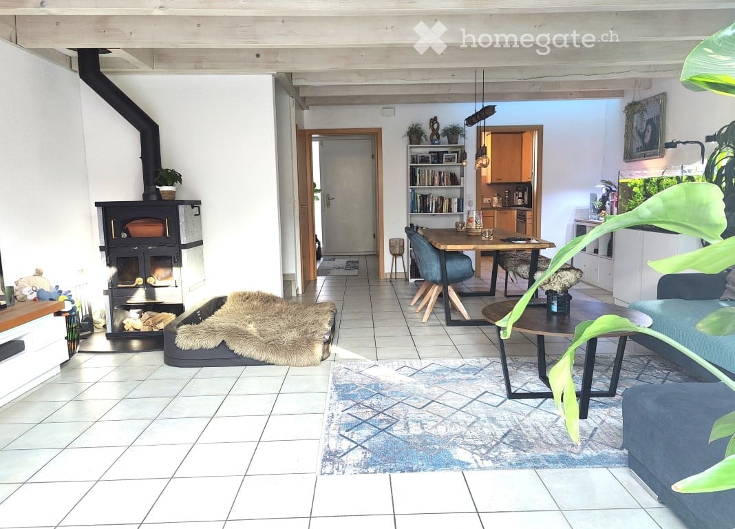 Bright, open-plan living and dining area with tiled flooring, a fireplace, a large sofa, coffee table, dining table with chairs, bookshelf, and decorative rug.