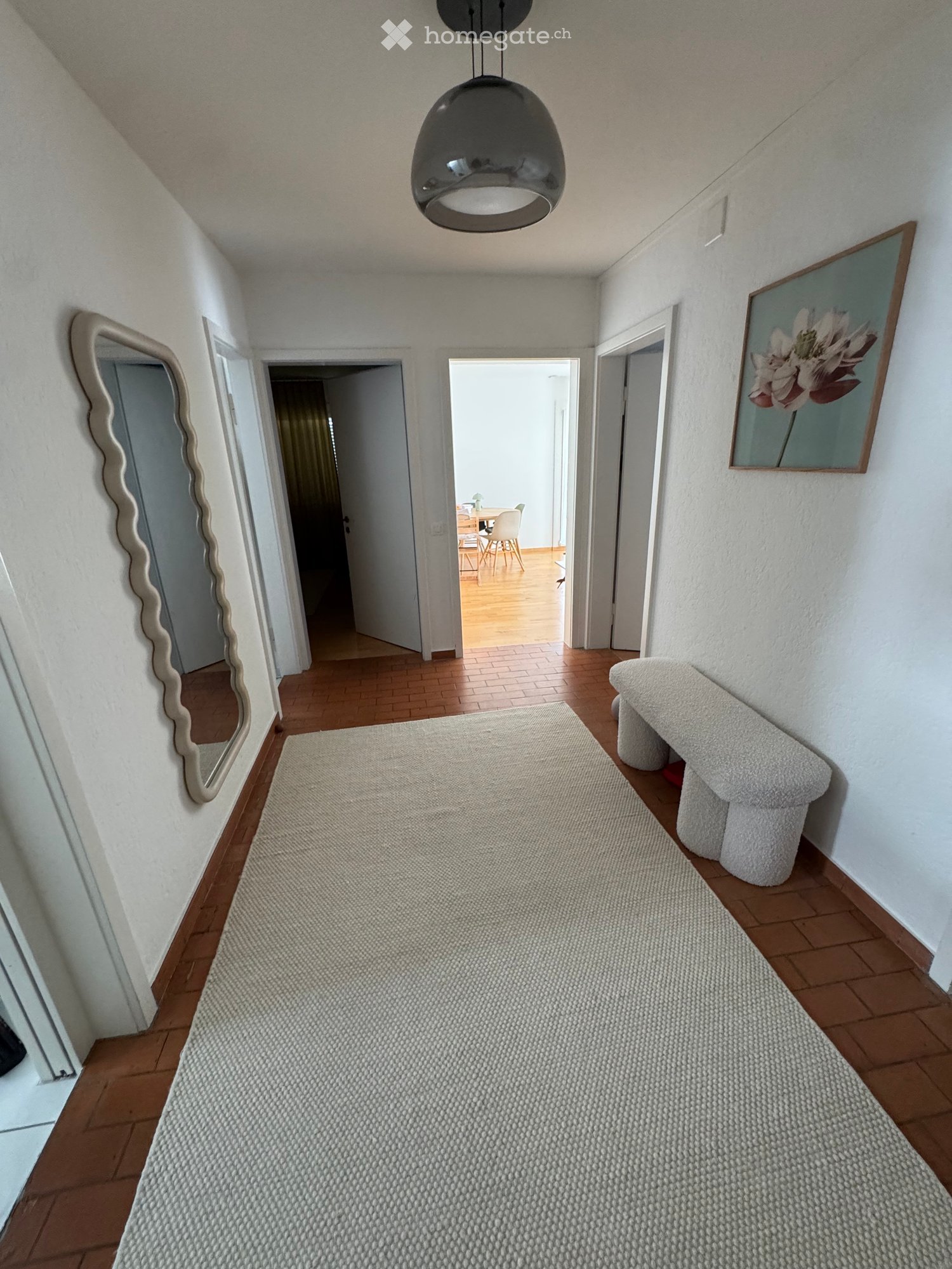 Hallway with wooden flooring, a large mirror, a bench, and a painting on the wall. The hallway leads to a dining area with a table and chairs.