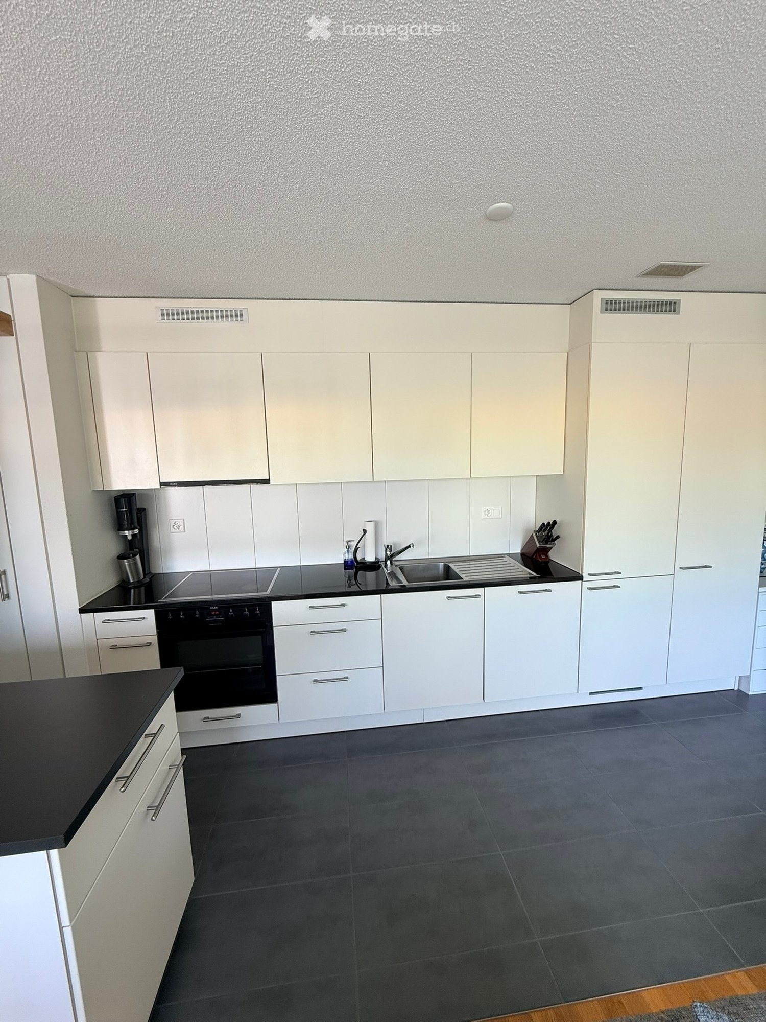 Modern kitchen with white cabinets, black countertops, and grey tiled floor. Features an oven, sink, faucet, coffee maker, and knife block.