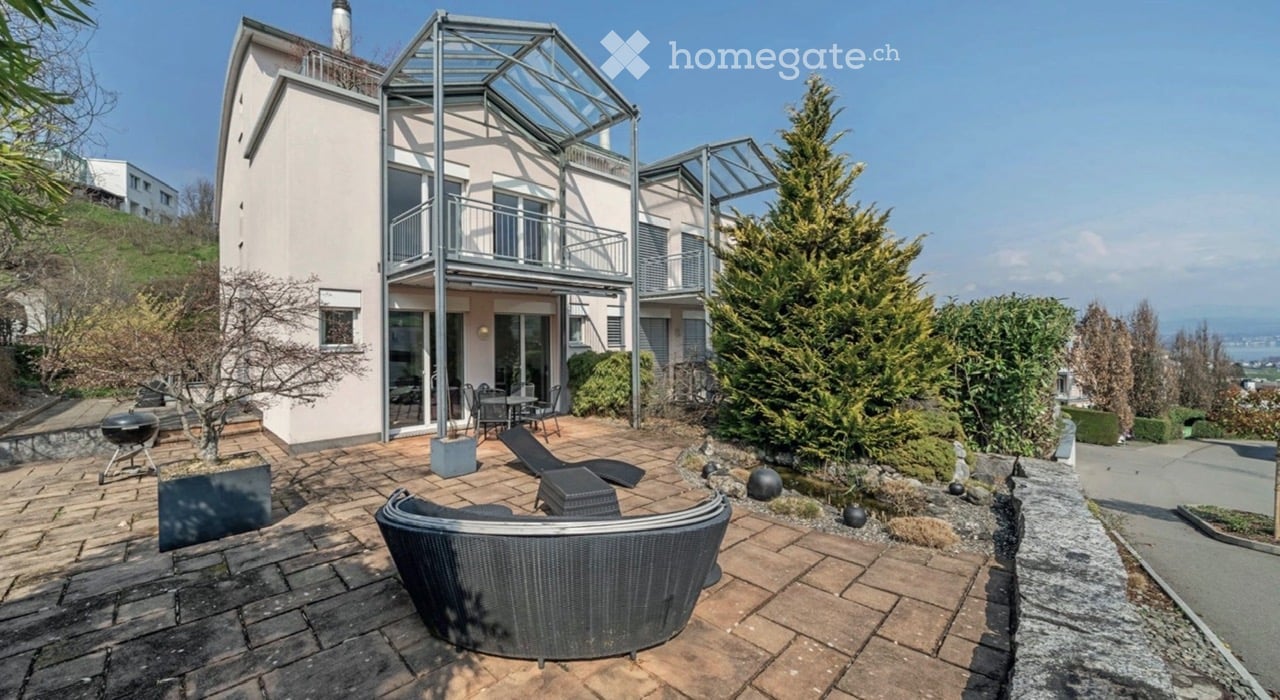 Modern outdoor patio with a wicker sofa, table, chairs, and a grill, adjacent to a two-story house with balconies and a glass roof structure.