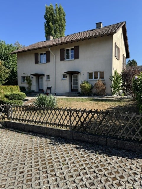 Two-story house with beige exterior, dark brown shutters, and a tiled roof. The house features a small porch with a door, multiple windows, and is surrounded by a garden with shrubs and trees. A wooden fence encloses the property.