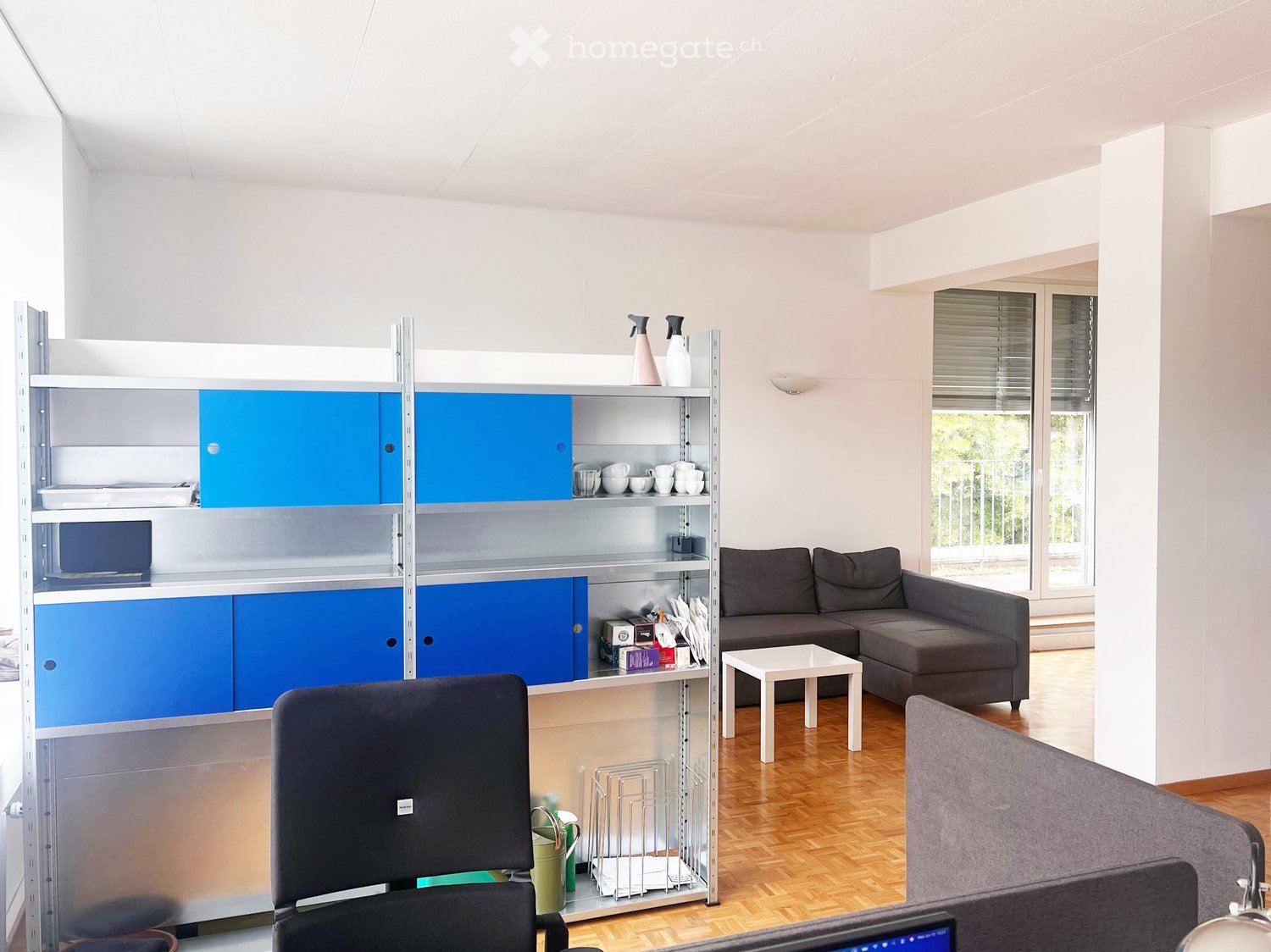 Modern living room with a grey sectional sofa, white coffee table, and a tall shelving unit with blue cabinets and various items on the shelves.