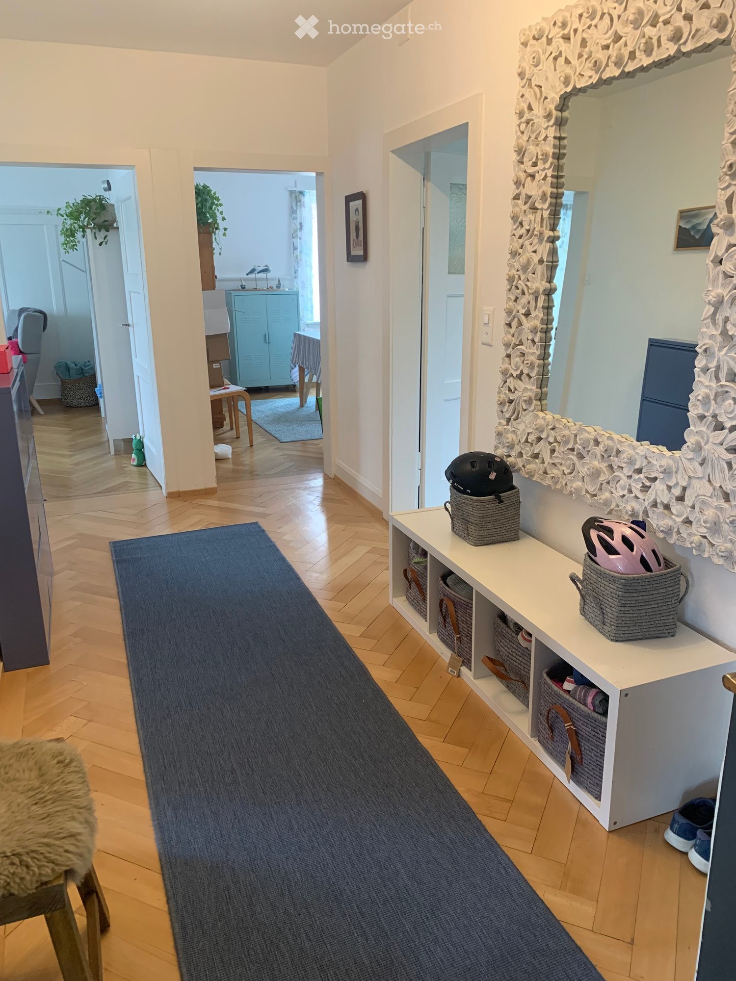 Hallway with wooden flooring, white walls, a large mirror with floral design, a white shelf with baskets, and a blue runner leading to another room.