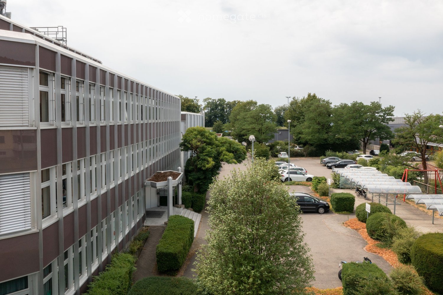 Modern multi-story building with a combination of red and grey facade, large windows, and a covered entrance. The surrounding area includes a parking lot with several cars, greenery, and a small playground.