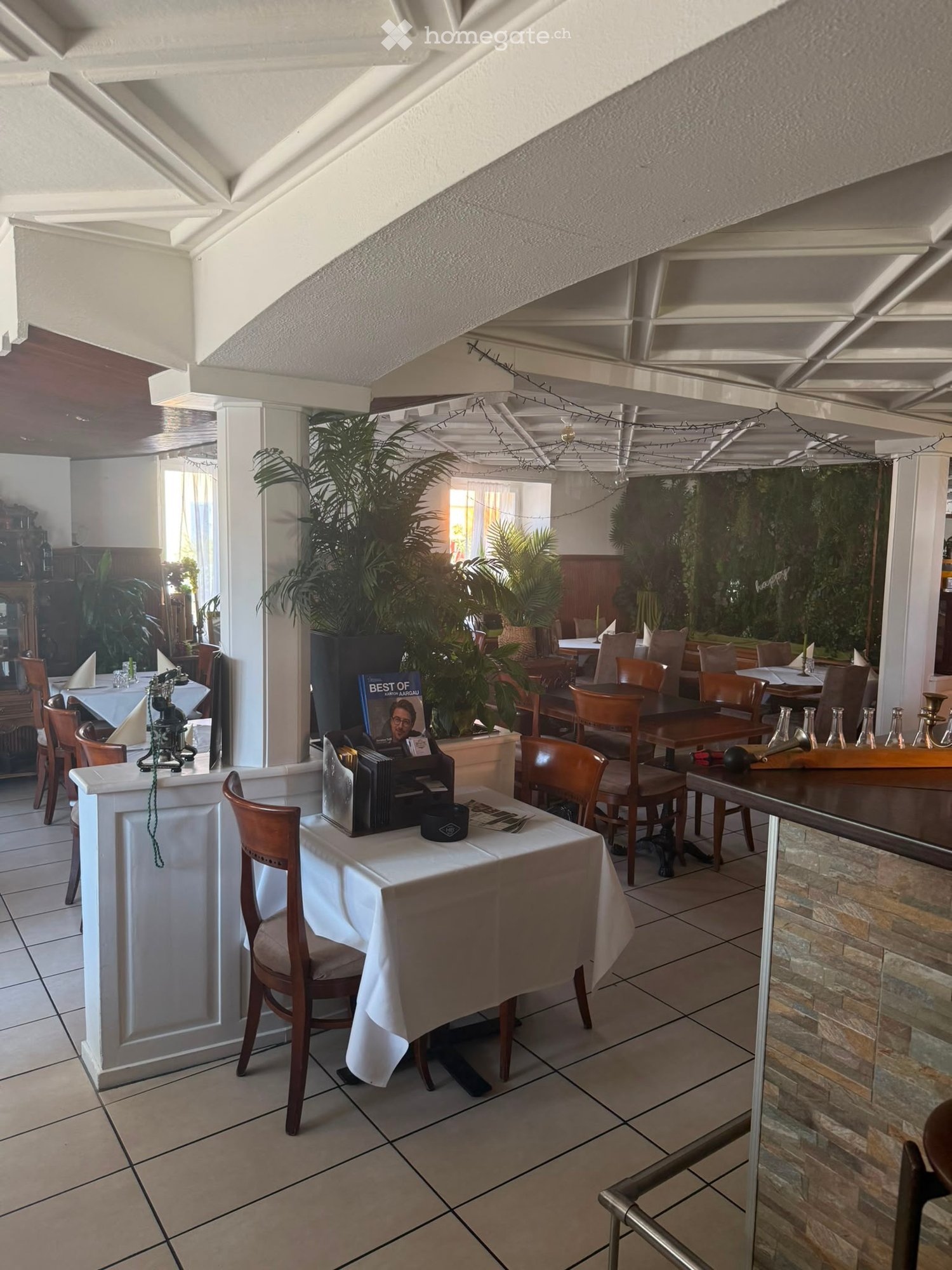 Restaurant interior with tiled floors, wooden tables, chairs, menus, and decorative plants.