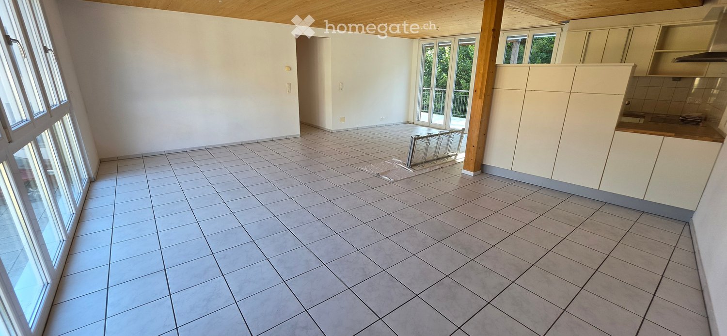 Empty kitchen with tiled floor, white cabinets, and large windows offering a view of greenery. No furniture or appliances are present.