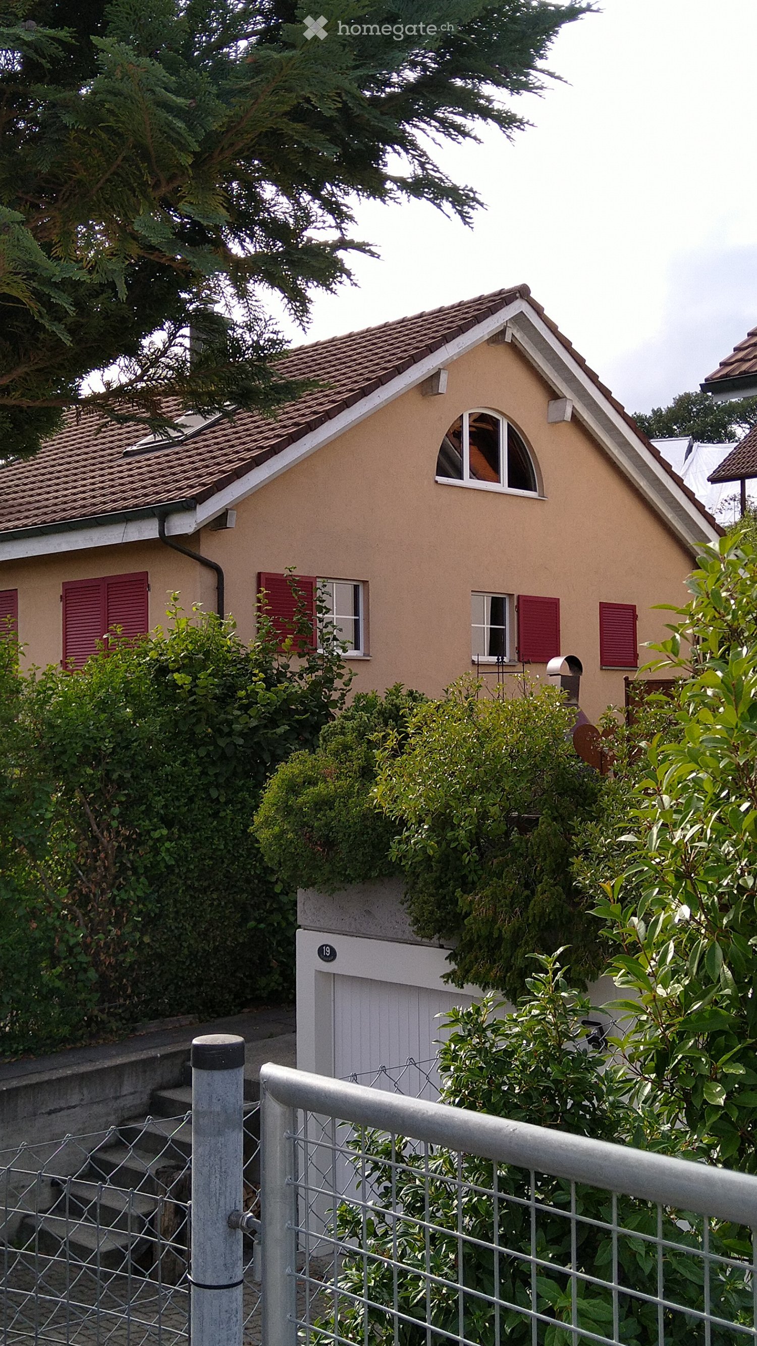A small, single-story house with a tiled roof, beige exterior, red shutters, and a white garage door. The house is surrounded by greenery and has a metal fence in the foreground.