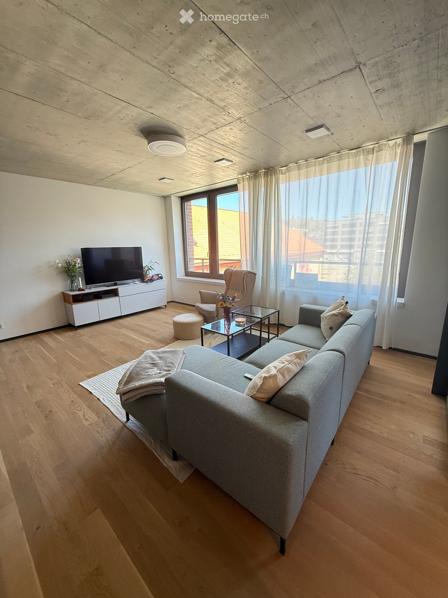 Modern living room with light wooden flooring, grey sectional sofa, coffee table, armchair, ottoman, TV stand, and decorative vases.