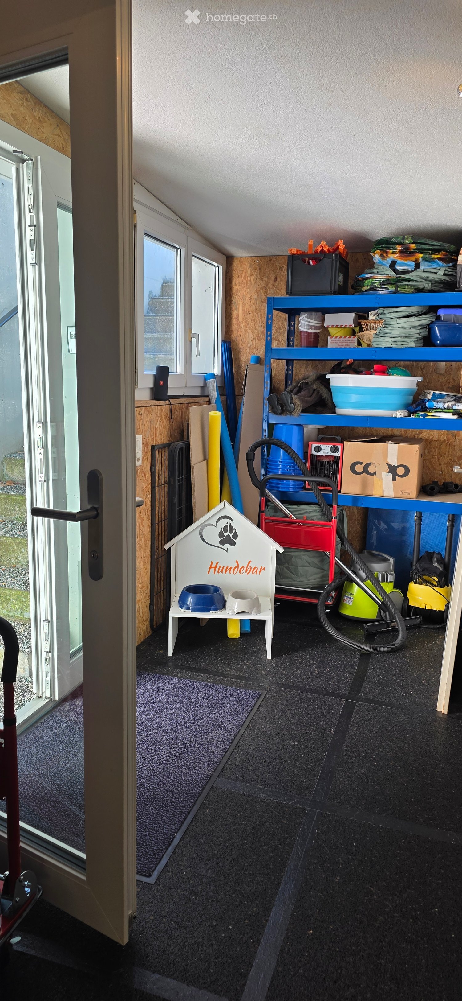 A well-organized storage or utility room with a blue shelving unit, vacuum cleaner, dog house with bowls, and various storage boxes.