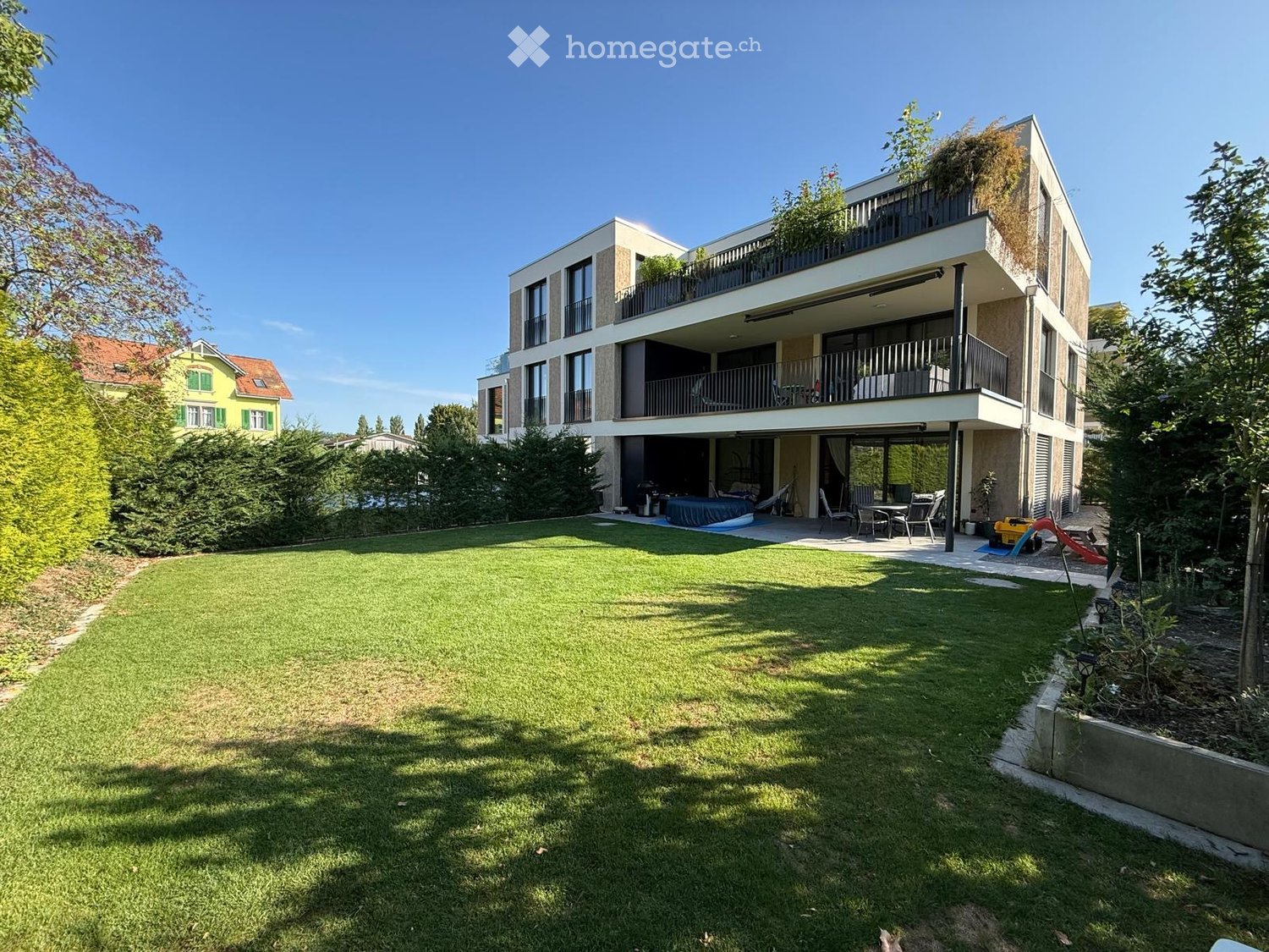 A modern two-story house with balconies, surrounded by a well-maintained lawn and garden area. The house features large windows and a contemporary design.