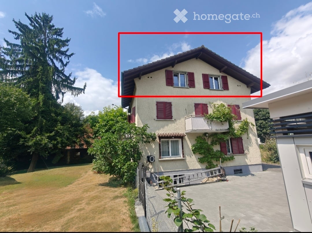 Two-story house with red shutters, white walls, and a balcony on the second floor. The house is surrounded by greenery and has a well-maintained lawn.
