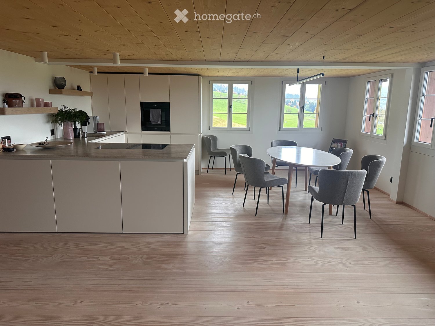 Open-plan kitchen and dining area with light wood flooring, white cabinets, and a central island. The dining area features a round table with six modern chairs.