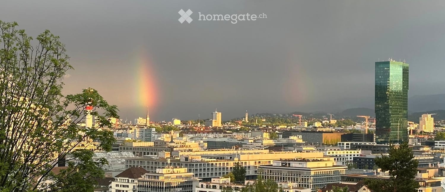 A panoramic view of a cityscape with a rainbow in the sky, featuring a mix of modern and older buildings, trees, and distant mountains.