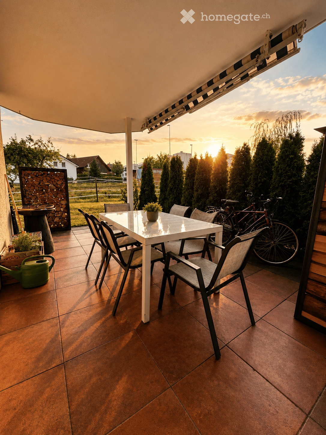 Balcony with a white table and chairs, bicycles, potted plants, and a garden hose, overlooking a suburban area at sunset.