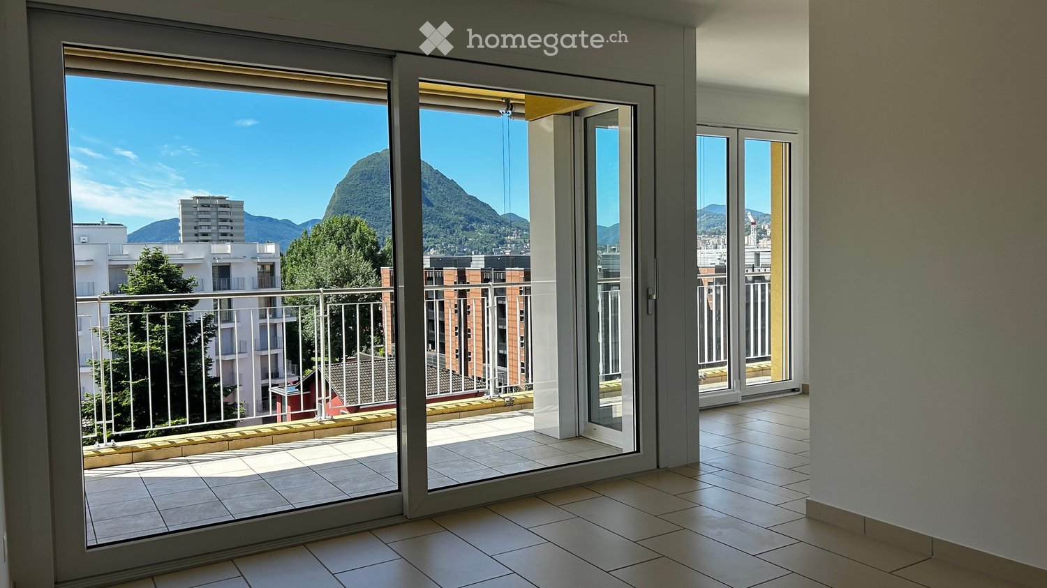 Open balcony with railing, large sliding glass doors, tiled floor, and distant city view with mountains.