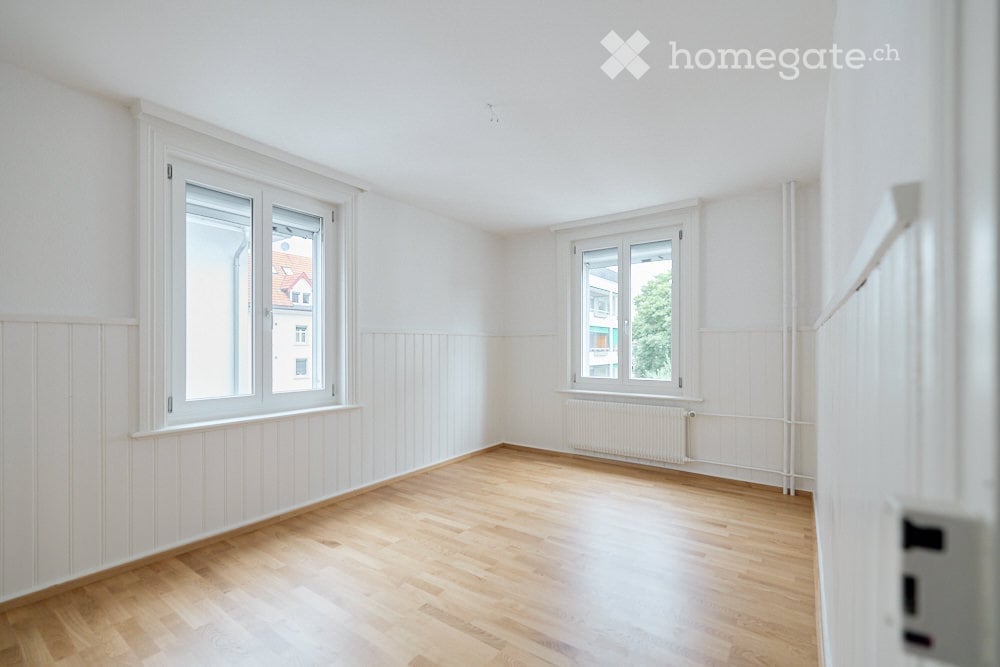 Completely empty room with bare white walls, wooden floor, and two windows with white frames. No furniture or equipment.
