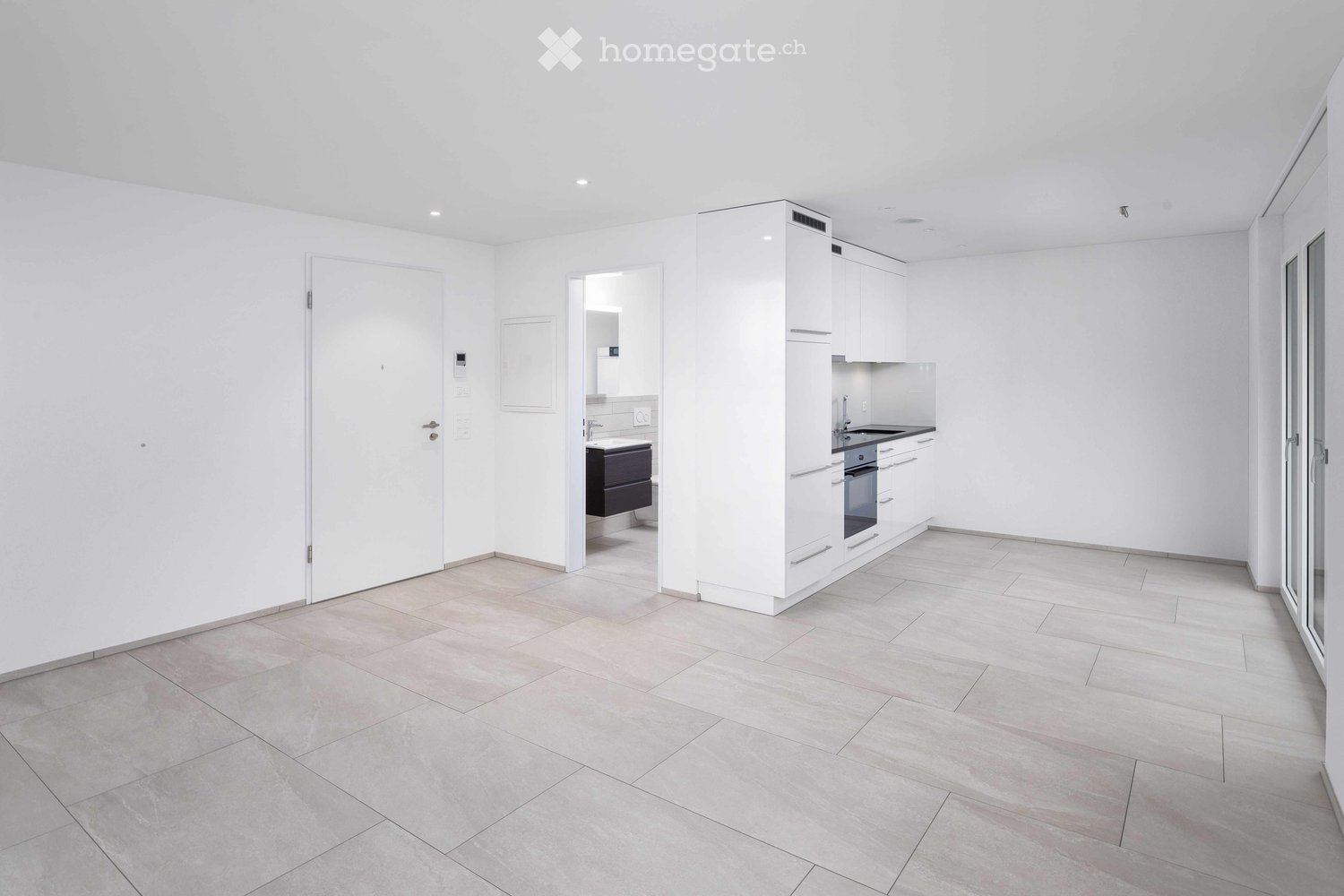 Empty room with white walls, tiled floor, and a doorway leading to a small bathroom area with a sink and dark cabinetry.