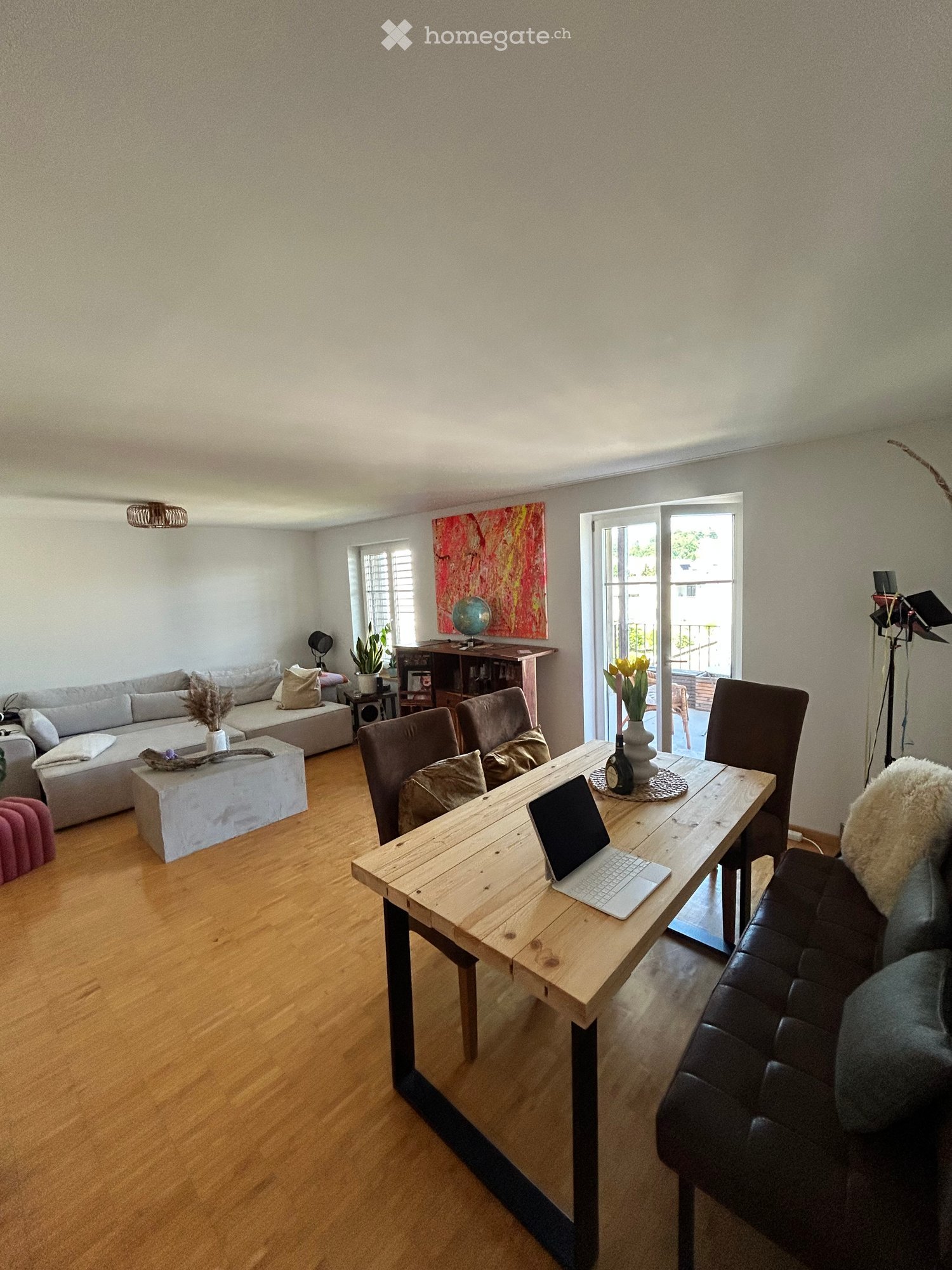 A modern living room with a wooden floor, a grey sofa, a coffee table, a laptop on a wooden table, and a leather armchair. There is a large window with a view of the balcony and a potted plant on the windowsill.