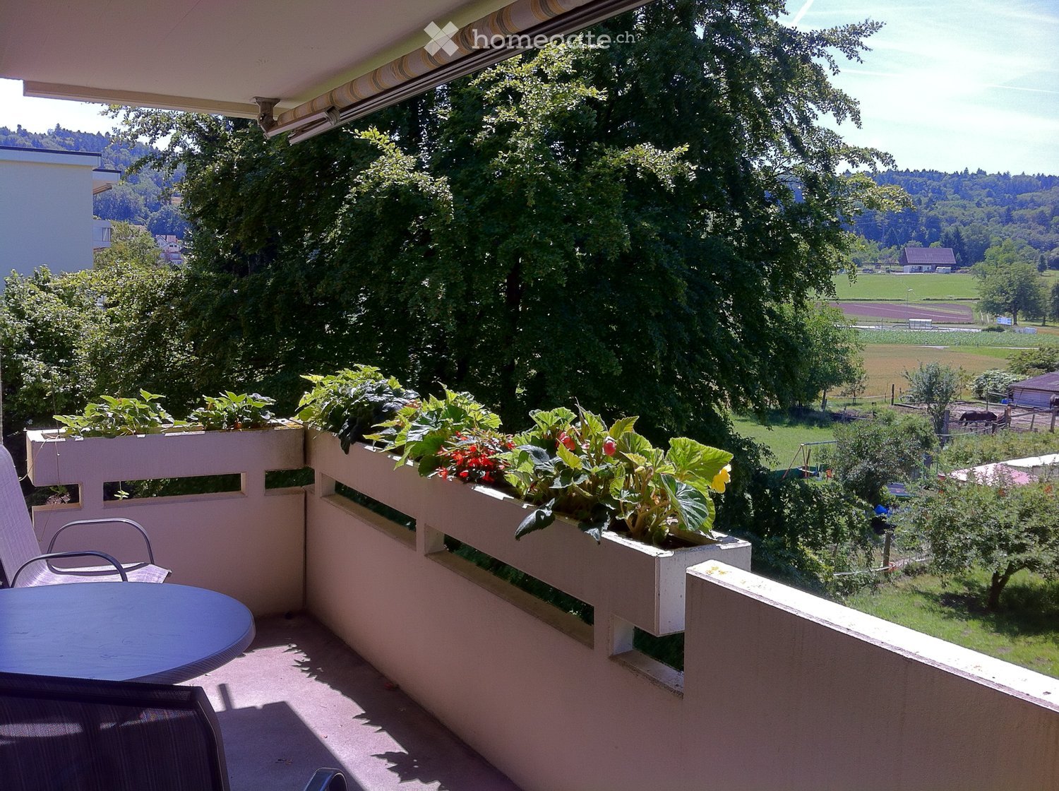 Balcony with a round table, chairs, and flower boxes, overlooking a scenic countryside view.