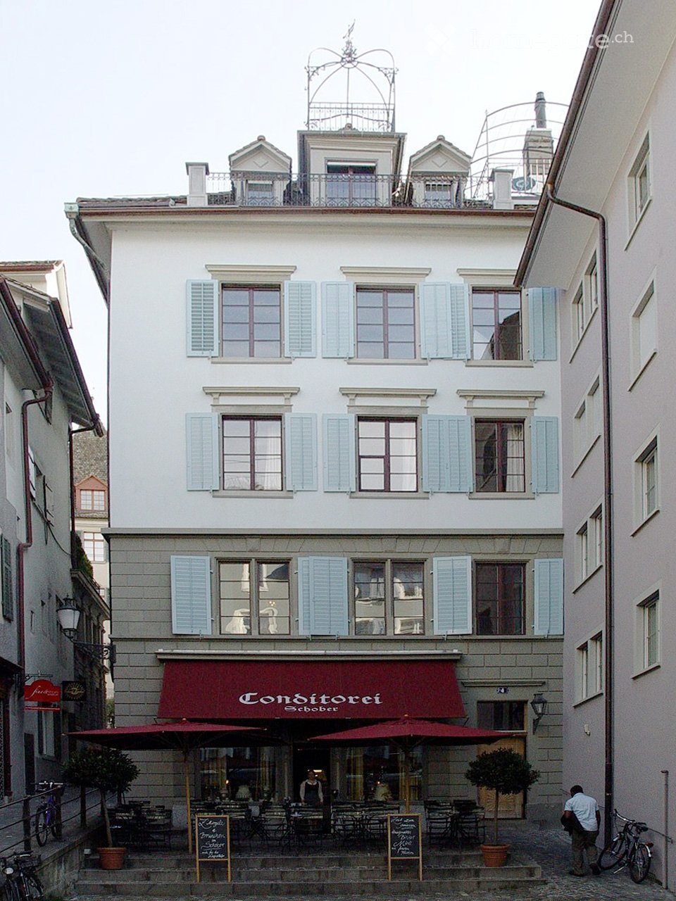 Three-story building with light-colored facade, multiple windows with shutters, and a red awning over the entrance. The ground floor features outdoor seating with tables and chairs under umbrellas.