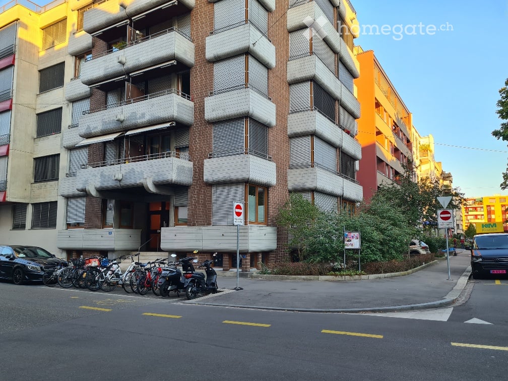 Multi-story apartment building with balconies, brick facade, and modern design. Several bicycles and scooters are parked along the sidewalk.