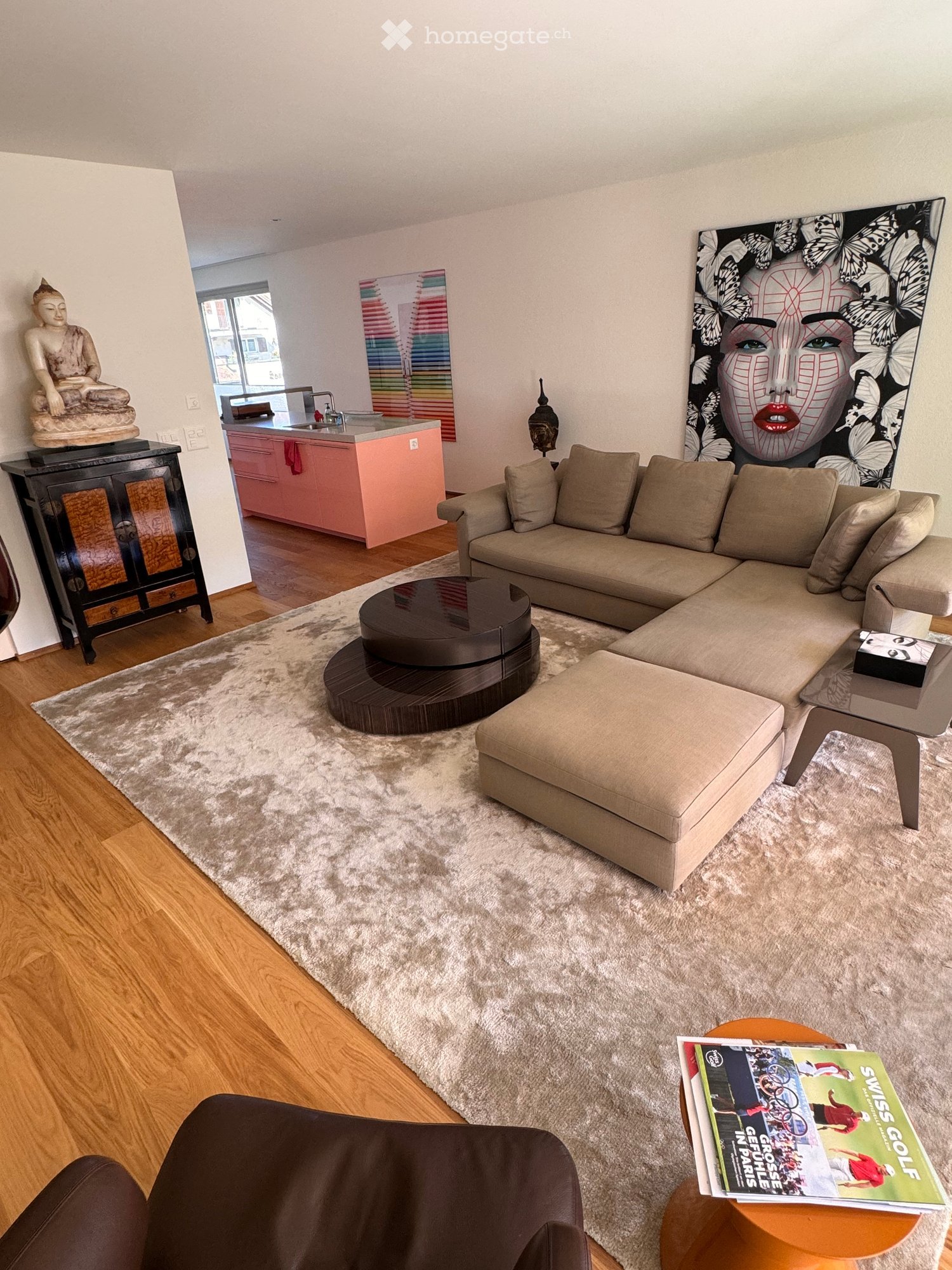 Modern living room with a large sectional sofa, ottoman, coffee table, and a small round table with magazines. Features a kitchen area with a pink counter, a black cabinet with a statue on top, and colorful artwork on the walls.