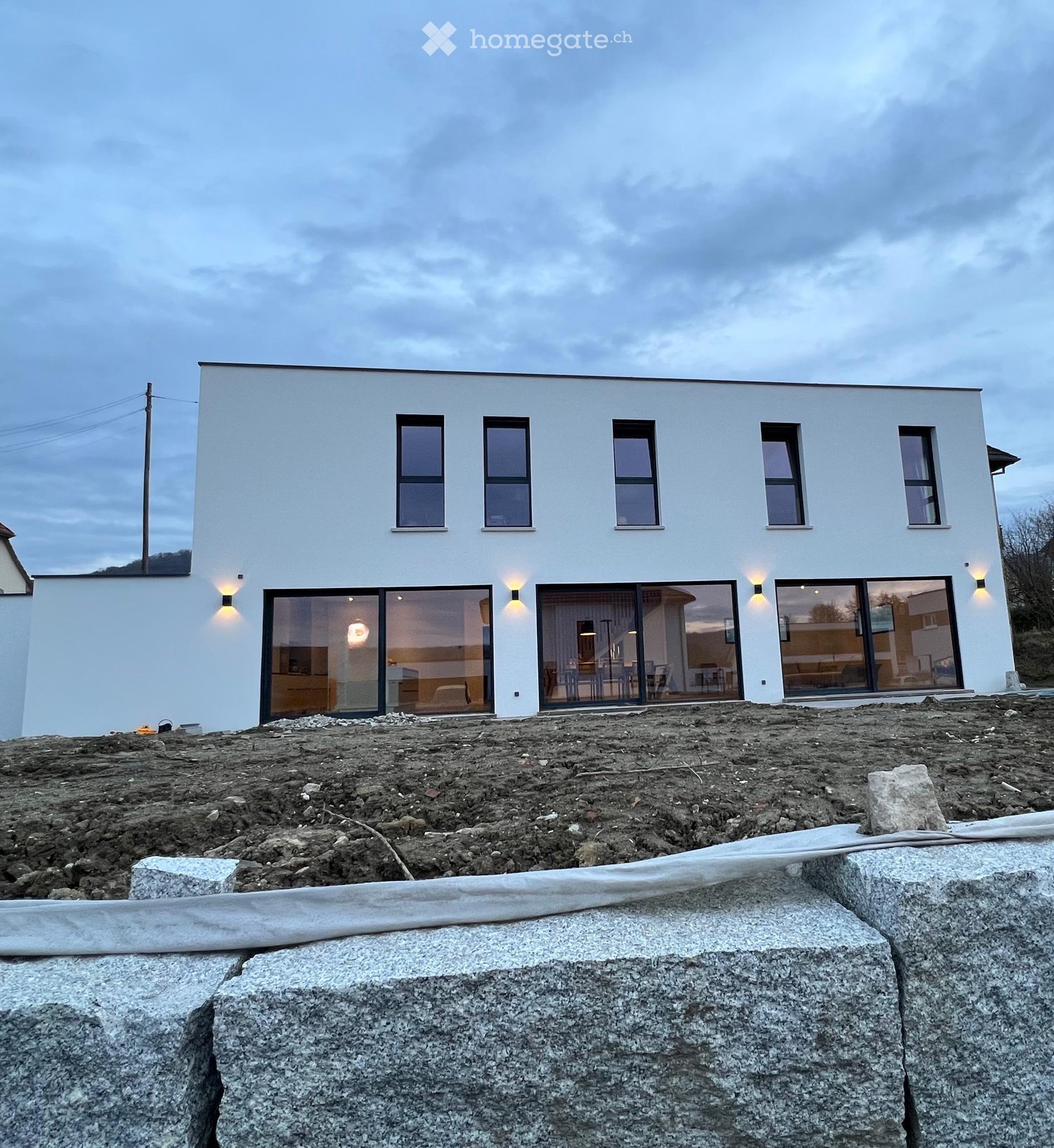 Modern single-story house with white exterior, large windows, and sliding glass doors, surrounded by construction materials and unfinished ground.