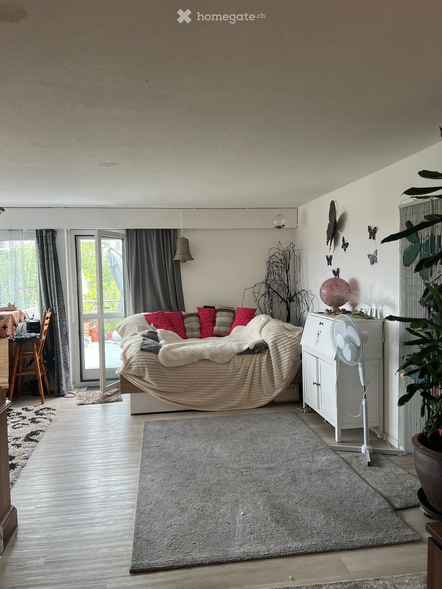 Cozy living room with a large sofa, colorful pillows, a fan, a small table with a chair, and decorative plants. The room features wooden flooring, a large window with curtains, and a gray rug.
