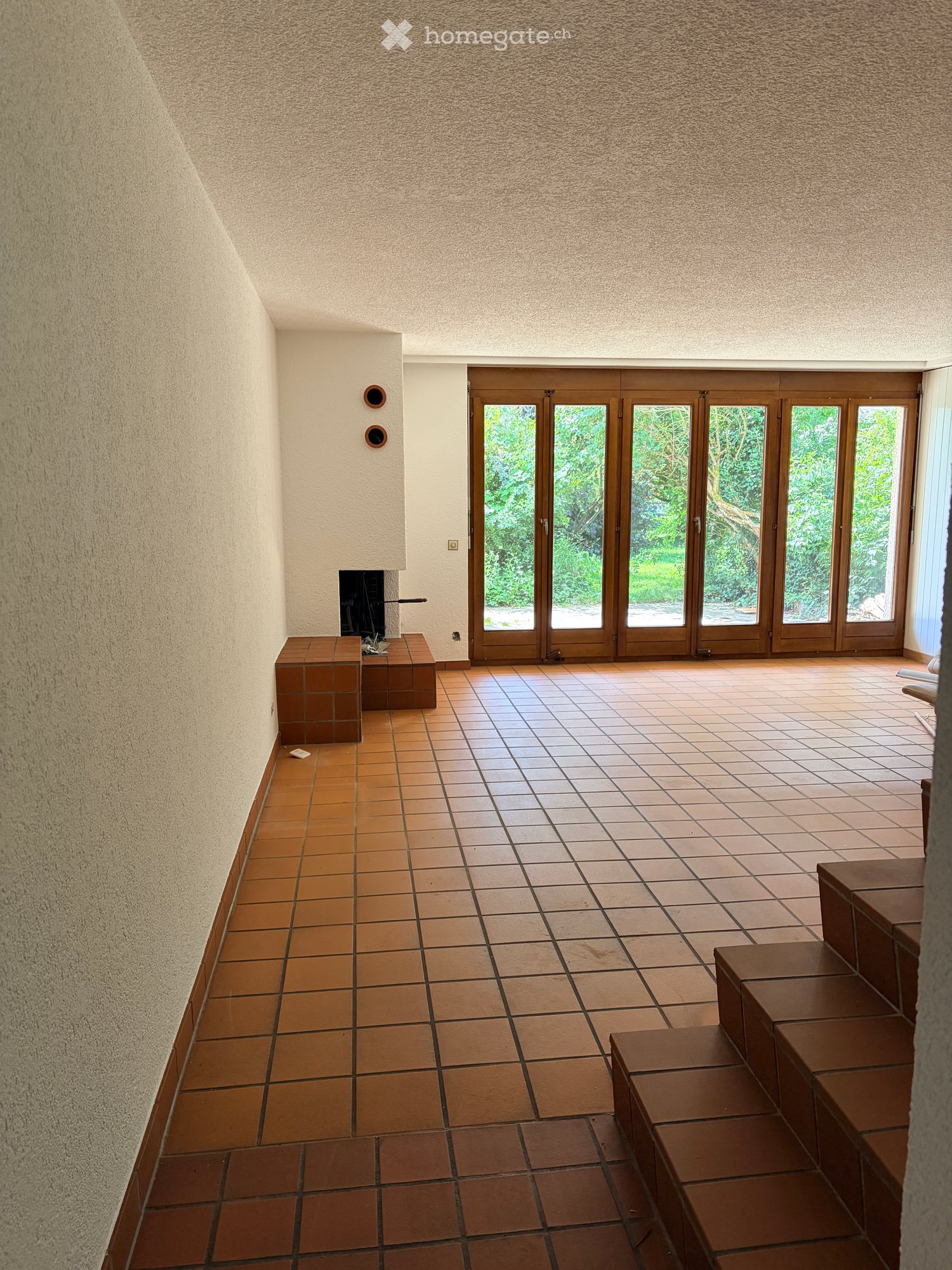 Empty hallway with tiled floor, textured walls, and a fireplace on the left side. Large glass doors lead to an outdoor area with greenery visible.