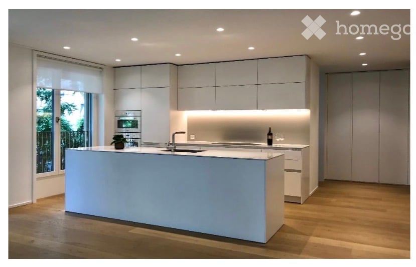 Modern kitchen with white cabinets, a central island, built-in oven, and sleek lighting. Features a large window with a view of greenery.